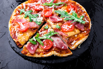 Homemade pizza with prosciutto, tomato, arugula on black slate board. Close up.