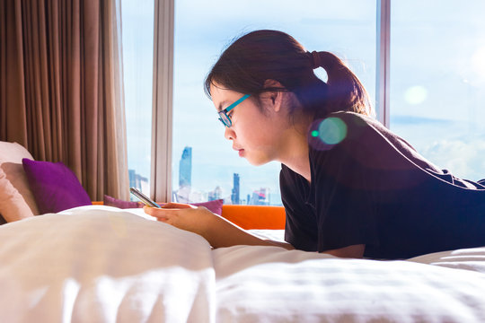 asian female teenager using her smart phone in bed, morning sunlight with flare