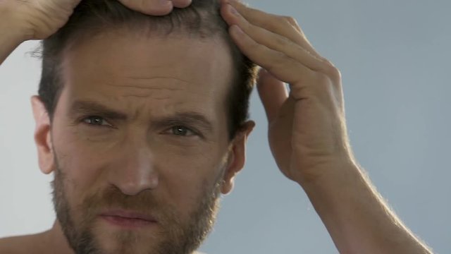 Bearded man standing in front of mirror and looking at his thinning hair, bald