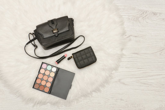 Black handbag, eye shadow, wallet and lipstick. White fur, top view. Fashionable concept