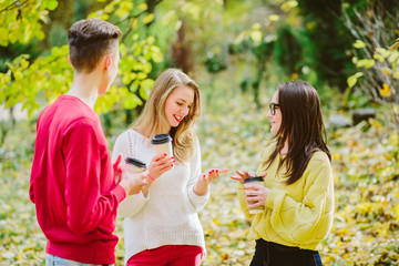 Two teenager girls and guy takling and drinking coffee or tea from disposable cup in green park. Education, lifestyle and people concept