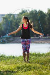 Sporty woman on the lakes coast makes exercise using a jump rope on the sunset