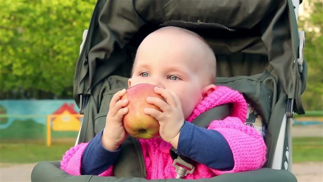 Baby girl sits in a baby stroller and eats an apple