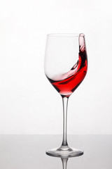Splashing and moving red wine in the wineglass standing against light background with reflection on the stand