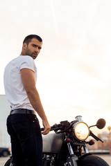 Sporty biker handsome rider man in white blank t-shirt walk to classic style cafe racer motorbike on rooftop at sunset. Vintage bike custom made in garage. Brutal urban lifestyle. Outdoor portrait.