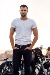 Fototapeta na wymiar Close up of a handsome rider man in white blank t-shirt look to camera near classic style cafe racer motorcycle at sunset. Bike custom made in vintage garage. Brutal urban lifestyle. Outdoor portrait.