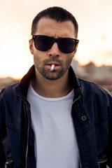 Obraz premium Close-up of a handsome rider guy with beard and mustache in black biker jacket, fashion sunglasses smoking cigaret near classic style cafe racer motorbike at sunset. Brutal urban lifestyle portrait.