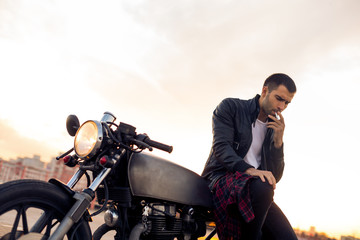 Fototapeta na wymiar Handsome rider man with beard and mustache in biker jacket smoking, thinking and sit on classic style cafe racer motorbike at sunset. Bike custom made in vintage garage. Brutal fun urban lifestyle.
