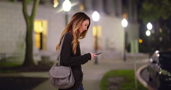 White female in her 20s messaging on cell phone, standing outside campus library at night. Caucasian millennial or student texting on smartphone outside building in the evening. 4k 