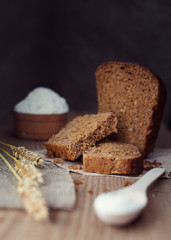 sliced brown bread with ears of wheat, flour or salt over dark wooden background, rustic style, close up