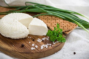 Closeup of round white homemade cheese - traditional milk creamy dairy product served with herbs and crackers and salt on vintage wooden board. Rustic style.