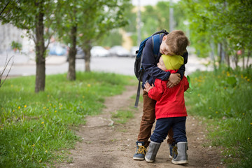 Two little brothers hugging each other when meeting in the park. Cute kid boy meets his little toddler sibling and hugs him. Happy children walking on the road. Lifestyle and family concept