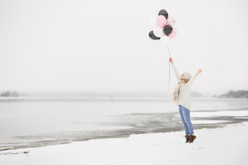 Happy young woman standing on a river shore and holding bunch of pink, silver, white and black ballons on a winter day. Birthday girl walking on the beach. Celebration, holidays.