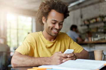 Cheerful handsome young African American male student in yellow t-shirt browsing internet on smart phone, having rest at coffee shop after classes at university, looking at screen and smiling broadly