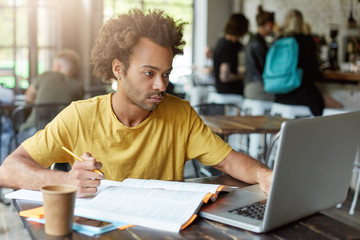 University student with dark skin and African hairstyle sitting at cafe working with books and...