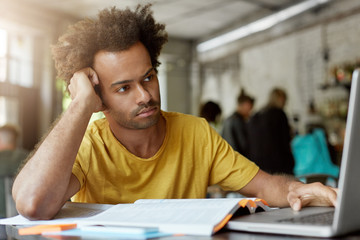 People, education, modern technologise and youth concept. Dark-skinned mixed race male with curly hair being focused in screen of laptop having thoughtful look leaning at his hand sitting in cafe