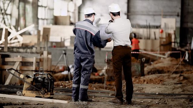 Back view of two men standing together in construction area and looking at shop drawings of future structure. Foreman in shirt and trousers is discussing with worker about details of building-up.