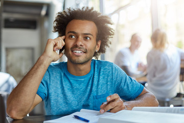 People, technology and communication concept. Handsome African American student with beard smiling, having nice phone conversation with his groupmate while working on home assignment at cafeteria