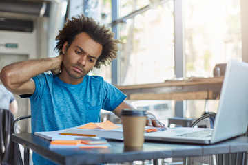 Serious Afro American male student in blue T-shirt sitting at cafeteria drinking takeaway coffee working at his project using books and laptop touching his neck with hand having pain after work