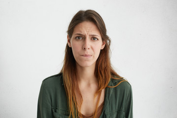 Sad young woman with long dark hair, charming eyes and pure skin curving her lips looking with disappointment having some troubles at work looking upset while posing in studio over white wall