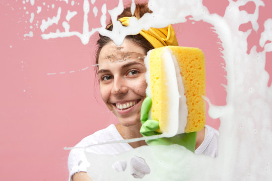 Hygiene, cleanliness, household chores, duties and domestic work concept. Beautiful housewife wearing green rubber gloves and headband, sponging window or mirror, wiping out foam off large glass