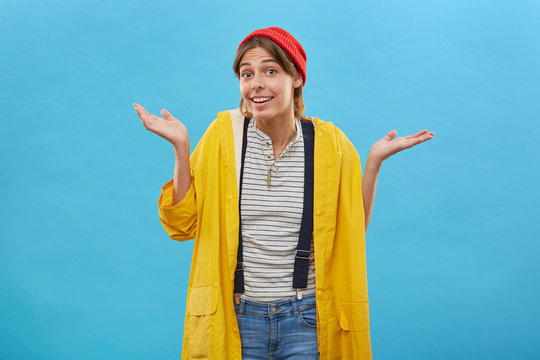 Nobody cares about it. Young pretty female wearing yellow anorak and jean overalls shrugging her shoulders having happy expression and hesitation, raising her eyebrows with uncertainty, gesturing
