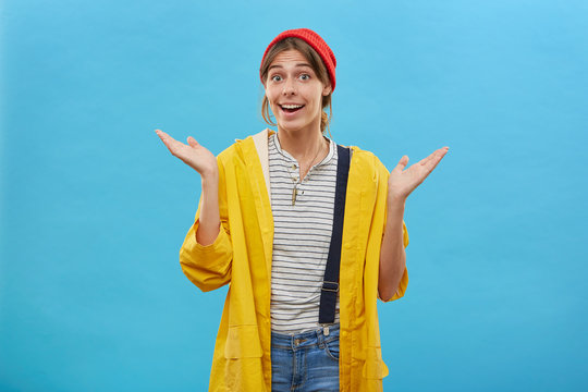 Beautiful housewife wearing red hat, yellow jacket and jean overalls shrugging her hands, raising eyebrows with surprisment, opening mouth feeling wonder posing over blue background with copy space