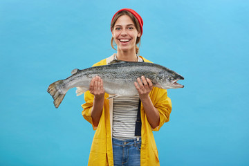 Glad fisher woman in yellow anorak and jeand overalls holding huge fish rejoicing to catch it demonstrating her work while standing over blue background. People, hobby, recreation and fishing