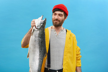 Portrait of glad fisherman wearing red hat, yellow jacket and overalls looking with pleased...
