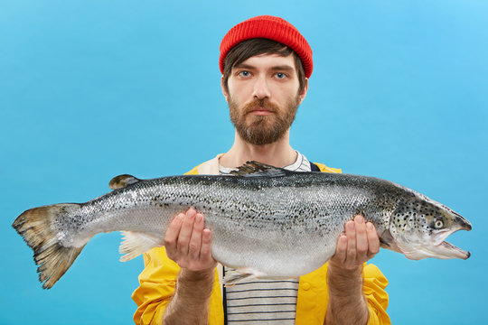 Horizontal portrait of successful angler with beard holding huge fish which he catched. Young fisherman dressed casually standing with huge trout. Man with fresh catch. Fishing and recreation
