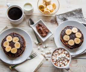 Breakfast for couple included oat pancakes with banana, nutella, honey, with coffe and milk and cacao with marshmallow on white wooden  background