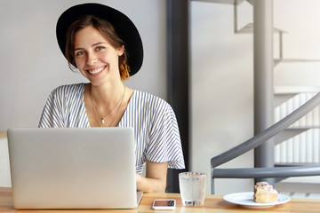 Businesswoman dressed officially sitting at her working place using laptop computer having pleasant smile rejoicing end of working day. Caucasian female wearing black hat having rest after work