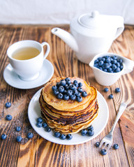 Pancakes healthy breakfast with blueberries, bog whortleberry, cup of green tea, cup of blueberries and teapot on brown wooden background 