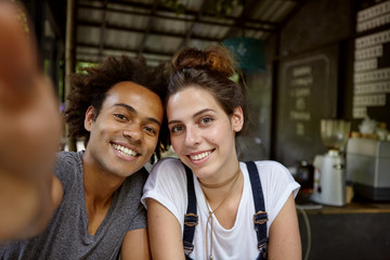 Glad mixed race male with curly hairstyle making photo with his female friend while resting with her at cafeteria. Cheerful interracial friends having fun together making selfie posing in camera