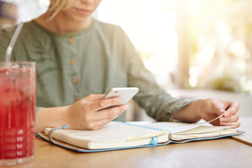 Cropped shot of young businesswoman sitting at cafe looking at her organiser and in calendar on smart phone looking for spare day to meet with her business partners, drinking red cold cocktail