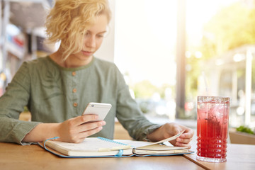 Cropped image of young blonde with trendy hairstyle sitting at cozy cafeteria drinking compote looking in her notebook holding cell phone calculating spendings. People, leisure, technology concept