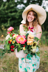wedding, floral design, beauty, country life concept - charming laughing woman in straw hat and summer dress with bouquet of grand beautiful peonies, soft yellow avalanches and bright red carnations