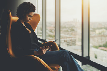 Cute pensive afro american student is sitting on armchair in front of window of skyscraper and having online conversation with her curator during probation period in office via digital tablet