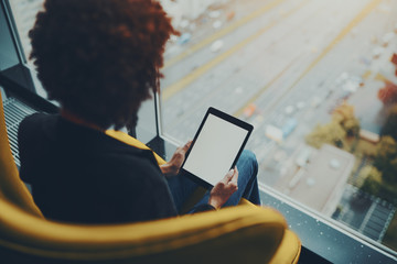 White mock-up of digital tablet screen in hands of black girl with afro hair sitting near window of skyscraper, blank template of digital pad screen in female hands of woman sitting in yellow armchair - Powered by Adobe