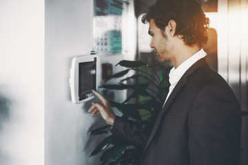 Fototapeta premium Handsome serious bearded employer in formal suit is adjusting lighting and air conditioning in office in the begging of work day using wall-mounted remote terminal with touch screen