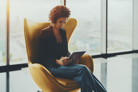Young cute Brazilian probationer using digital tablet to prepare for interview while sitting in yellow armchair, pensive afro american girl working on digital pad in office settings near huge window - Powered by Adobe