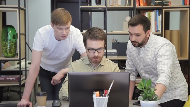 Three serious programmers brainstorm before the laptop. A man with a beard sits in front of the monitor and quickly prints on the keyboard. Two young guys stand on each side and point to important