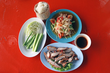 Thai Northeastern food set, which consists of papaya salad, Grilled Pork and sauce, Sticky rice in the wicker and Vegetable (cabbage, Long bean, cucumber) on the red table.