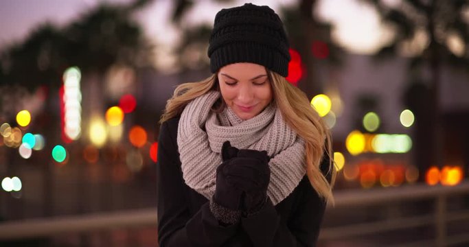 Attractive millennial girl wearing beanie, scarf, and gloves, blowing into her hands downtown in the evening. Young Caucasian woman in winter wear, standing outside on city street at night. 4k 