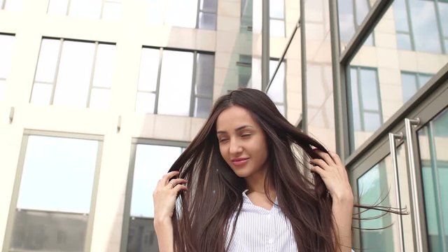 Close-up of a sexy girl with long dark hair on the background of modern glass building. Girl is holding her long hair with her hands.