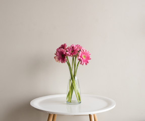 Pink gerberas in glass vase on small white round table against neutral wall background