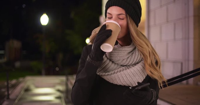 Millennial Girl In Winter Wear Drinking Coffee Or Tea And Laughing At Message On Her Phone Outside Building. Young Caucasian Woman Rolling Her Eyes At Her Cellphone And Smiling Downtown At Night. 4k 