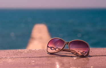 Glasses on the seaside. View of sunglasses on the beach near the sea with sand. Pink sunglass on terrace at seaside with nice blue water. Picture with copy space