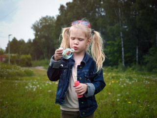 Obraz premium Girl inflates soap bubble. Portrait of a child. Long blonde hair, beautiful face. Nature, field, green, grass, forest