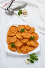 Baked sweet potatoes in white plate on a white wooden table with parsley, garlic and salt. White wooden rustic picnic table scenery from above.